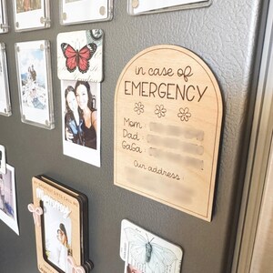 May include: A wooden sign with rounded edges and a white background. The sign says "in case of EMERGENCY" with a space for names and an address. The sign is attached to a refrigerator with magnets.