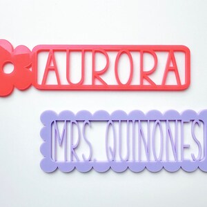 May include: Two personalised name tags, one with a pink flower and the name "AURORA" in white, and the other with a scalloped border and the name "MRS. QUINONES" in white.