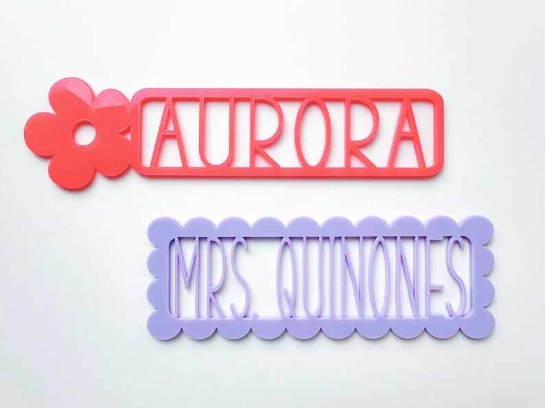May include: Two personalised name tags, one with a pink flower and the name "AURORA" in white, and the other with a scalloped border and the name "MRS. QUINONES" in white.