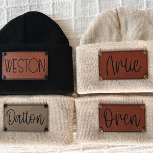 May include: Four knit beanies with leather patches. The beanies are in two colours: black and beige. The patches have the names Weston, Arlie, Dalton, and Oren.