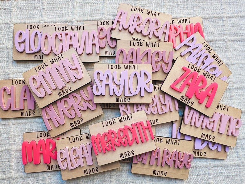 May include: A collection of personalized wooden name plaques. Each plaque features a name in a layered, bubble-style font, with a contrasting color. The text "LOOK WHAT MADE" is also included on each plaque.