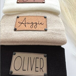 May include: Three knitted beanies with personalised leather patches. The top beanie is white with a brown leather patch that says "Cade". The middle beanie is beige with a brown leather patch that says "Auggie". The bottom beanie is black with a tan leather patch that says "Oliver".