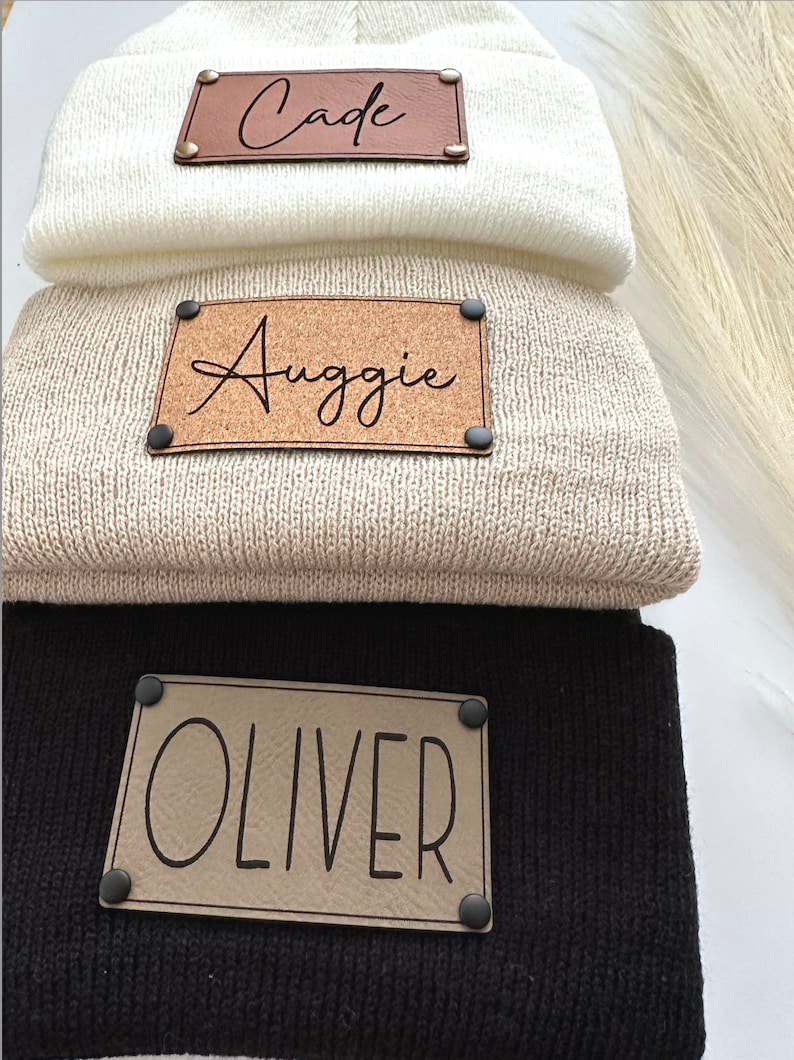 May include: Three knitted beanies with personalised leather patches. The top beanie is white with a brown leather patch that says "Cade". The middle beanie is beige with a brown leather patch that says "Auggie". The bottom beanie is black with a tan leather patch that says "Oliver".