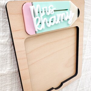 May include: A wooden frame with a cutout in the shape of a pencil. The pencil is painted light blue and pink and has the words "Mrs. Khamy" written on it in white.