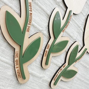 K&ouml;nnte beinhalten: Florale Holzausschnitte mit gr&uuml;nen Blattakzenten. Der Text "Happy Mothers Day!" und Namen sind in das Holz graviert. Die Ausschnitte sind mit einer hellen Holzbasis und einem dunkleren Holzrand geschichtet.