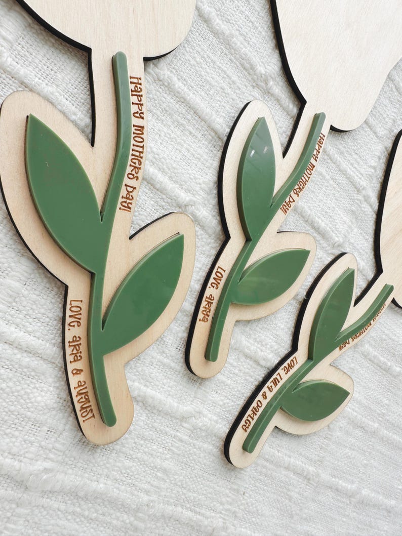 K&ouml;nnte beinhalten: Florale Holzausschnitte mit gr&uuml;nen Blattakzenten. Der Text "Happy Mothers Day!" und Namen sind in das Holz graviert. Die Ausschnitte sind mit einer hellen Holzbasis und einem dunkleren Holzrand geschichtet.
