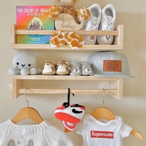 May include: A wooden shelf unit with two shelves and a hanging rod. The top shelf has a stuffed giraffe toy, a book titled "The Colors of Snow", and a wooden sign that says "oh baby". The middle shelf has a crocheted octopus, a pair of baby shoes, and a gray baseball cap with the name "AIDEN" on it. The bottom shelf has a white knitted sweater with the name "Aiden" on it, a pair of red and white crocheted baby shoes, and a white onesie with the word "Supercute" printed on it.