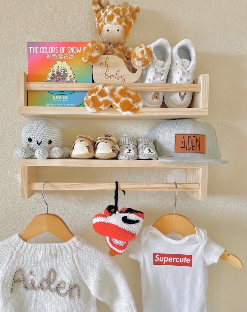 May include: A wooden shelf unit with two shelves and a hanging rod. The top shelf has a stuffed giraffe toy, a book titled "The Colors of Snow", and a wooden sign that says "oh baby". The middle shelf has a crocheted octopus, a pair of baby shoes, and a gray baseball cap with the name "AIDEN" on it. The bottom shelf has a white knitted sweater with the name "Aiden" on it, a pair of red and white crocheted baby shoes, and a white onesie with the word "Supercute" printed on it.