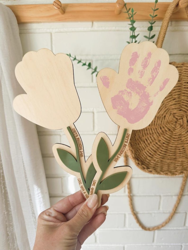 K&ouml;nnte beinhalten: Holzblumenf&ouml;rmiges Muttertagsgeschenk mit einem Handabdruck in rosa Farbe. Die Blume hat gr&uuml;ne Bl&auml;tter und Stiele mit dem Text "Happy Mother's Day" und "Love you always".