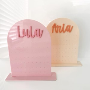 May include: Two earring display stands. One is pink with the name "Lula" in cursive, and the other is peach with the name "Aria". Both have a series of small holes for holding earrings.