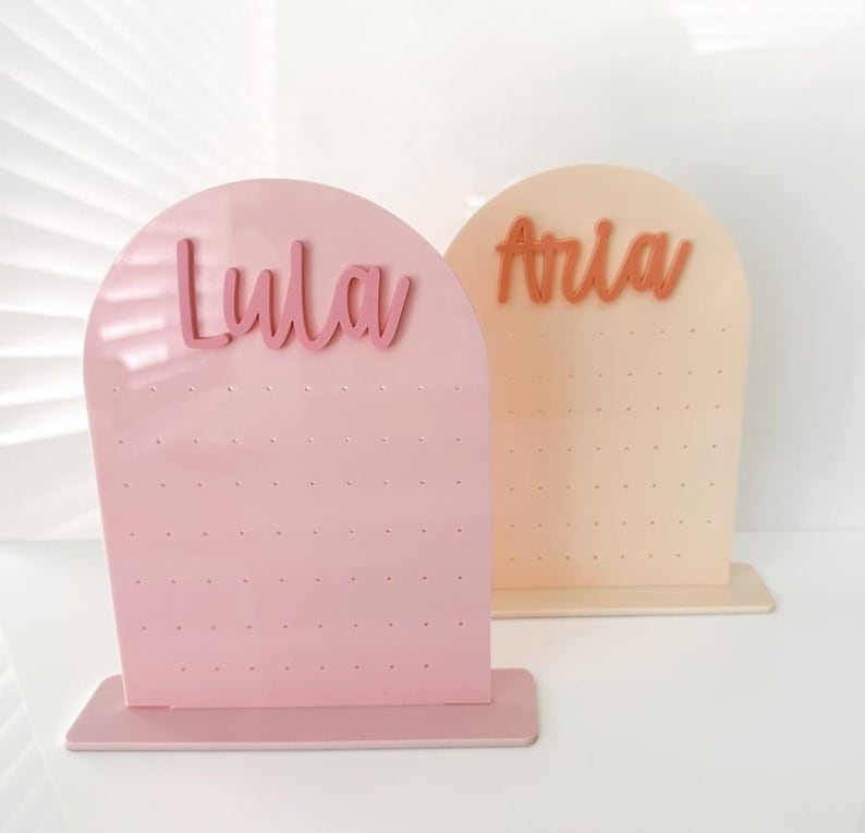 May include: Two earring display stands. One is pink with the name "Lula" in cursive, and the other is peach with the name "Aria". Both have a series of small holes for holding earrings.