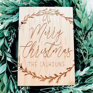 Pu&ograve; includere: Un cartello di legno con un design a corona di Natale e il testo "Merry Christmas The Calhouns".
