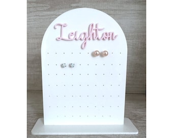 May include: White wooden earring holder with the name Leighton in pink lettering. The holder has multiple holes for earrings and features a pink pearl earring and a pair of silver earrings.