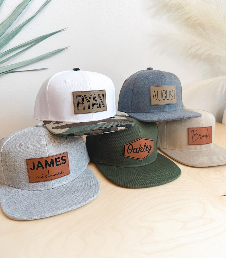 May include: Five different baseball caps with personalized leather patches. The caps are in various colors, including white, gray, green, and beige. The patches feature the names "Ryan", "August", "James Michael", "Oakley", and "Brooks".