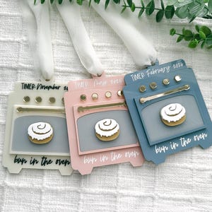 May include: Three oven-shaped ornaments in cream, pink, and blue, each with a cinnamon roll design. The ornaments have the text "TIMER" and "bun in the oven" with different months and years. They are hanging from white ribbons.
