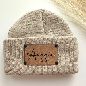 May include: A beige knitted beanie with a cork patch that says "Auggie" in black cursive.