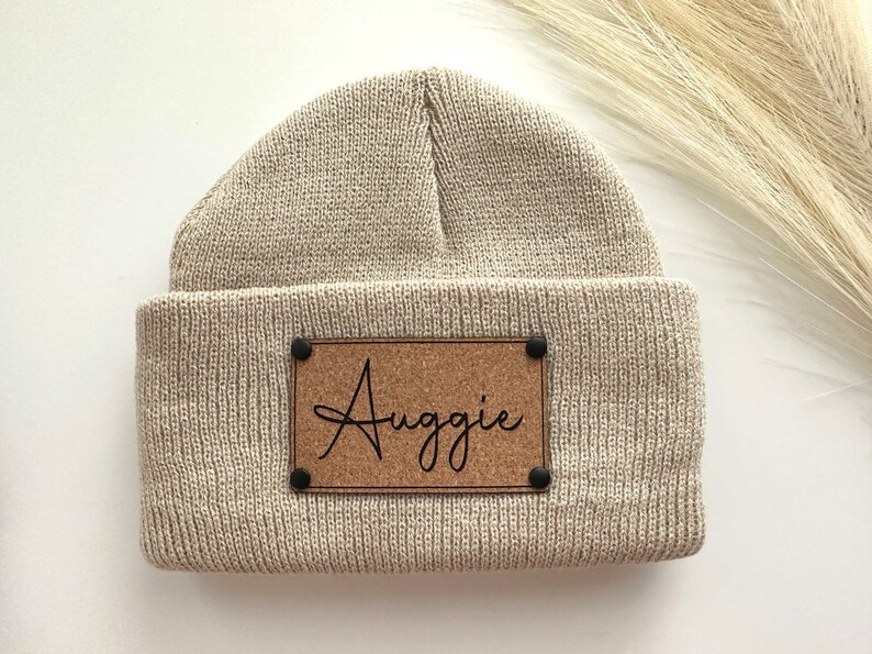 May include: A beige knitted beanie with a cork patch that says "Auggie" in black cursive.