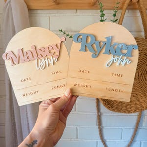May include: Two wooden arch-shaped signs with the names "Maisey Lynn" and "Ryker John" in pink and blue lettering. Each sign has spaces to record the date, time, weight, and length.