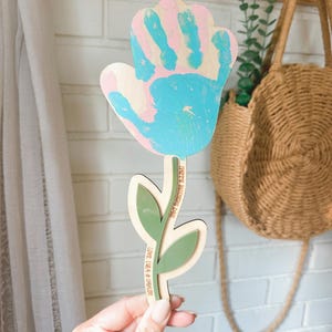 K&ouml;nnte beinhalten: Blumenf&ouml;rmiges Holzhandwerk mit einem Handabdruck-Design in rosa und blauer Farbe. Der Stiel hat gr&uuml;ne Bl&auml;tter und den Text "HAPPY MOTHER'S DAY" und "LOVE LILA & ORACLE". Ein dekorativer Artikel f&uuml;r den Muttertag.