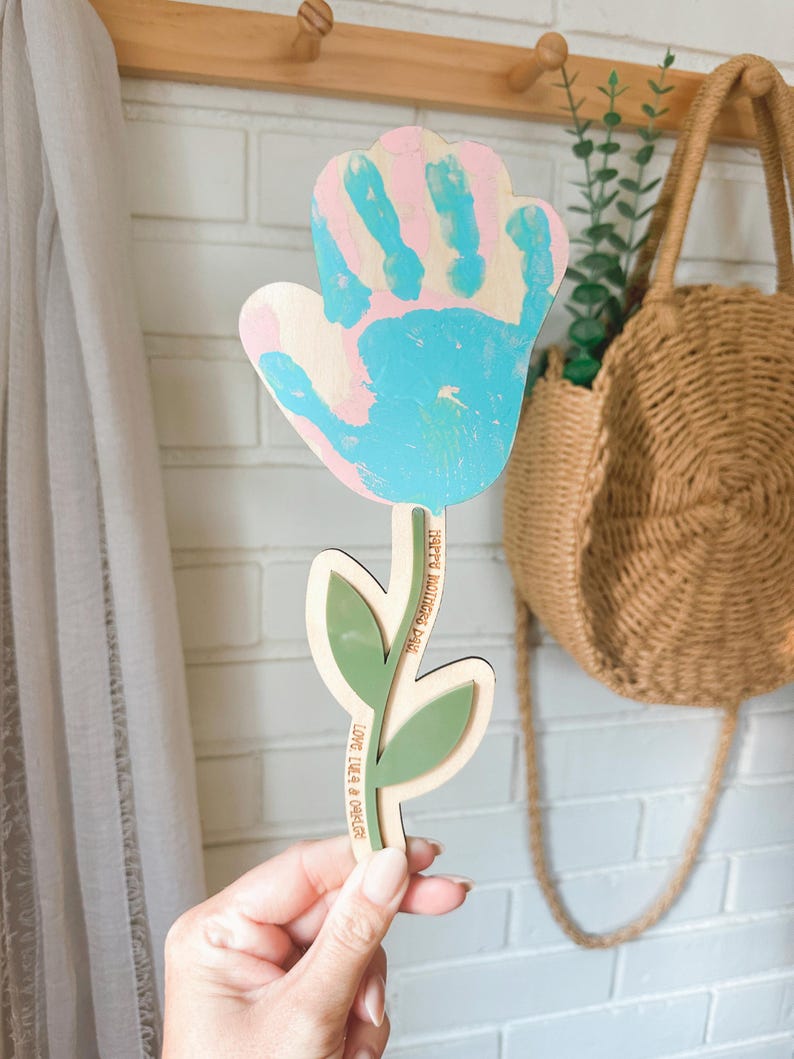 K&ouml;nnte beinhalten: Blumenf&ouml;rmiges Holzhandwerk mit einem Handabdruck-Design in rosa und blauer Farbe. Der Stiel hat gr&uuml;ne Bl&auml;tter und den Text "HAPPY MOTHER'S DAY" und "LOVE LILA & ORACLE". Ein dekorativer Artikel f&uuml;r den Muttertag.