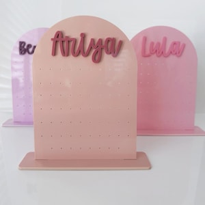 May include: Three pastel-coloured earring display stands. The centre stand is a light pink with the name "Ariya" in a darker pink script. The other two stands are lavender and pink, with the names "Ber" and "Lula" in script.
