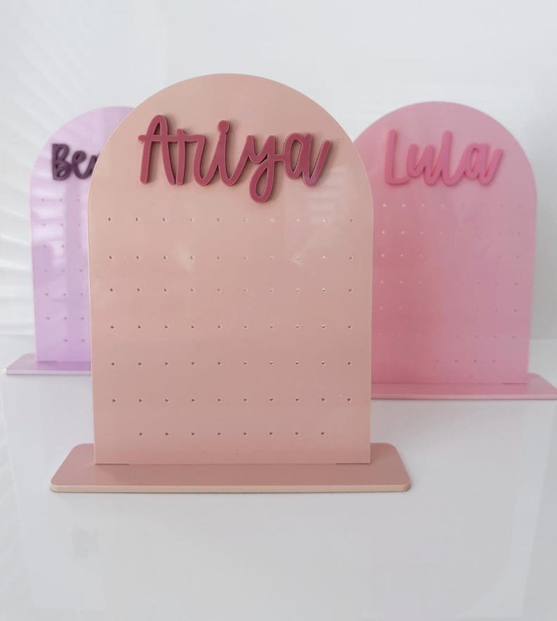 May include: Three pastel-coloured earring display stands. The centre stand is a light pink with the name "Ariya" in a darker pink script. The other two stands are lavender and pink, with the names "Ber" and "Lula" in script.