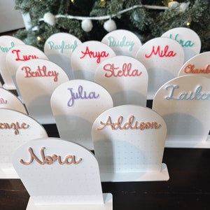 May include: White display stands with names written in various colours, including pink, blue, green, and orange. The names include Addison, Nora, Julia, Bentley, Stella, Aspen, Kenzie, Rayleigh, Arya, Paisley, Mila, Ira, Allie, Chamberlain, and Lauken.