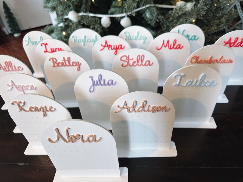 May include: White display stands with names written in various colours, including pink, blue, green, and orange. The names include Addison, Nora, Julia, Bentley, Stella, Aspen, Kenzie, Rayleigh, Arya, Paisley, Mila, Ira, Allie, Chamberlain, and Lauken.