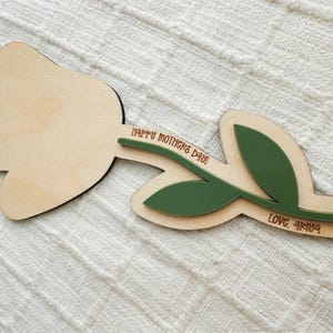 K&ouml;nnte beinhalten: Holzdekoration in Tulpenform mit hellem Holzfinish. Der Stiel und die Bl&auml;tter sind gr&uuml;n. Der Text "HAPPY MOTHERS DAY!" und "LOVE, ARYA" sind auf dem Stiel eingraviert. Die Dekoration liegt auf einer wei&szlig;en, strukturierten Oberfl&auml;che.
