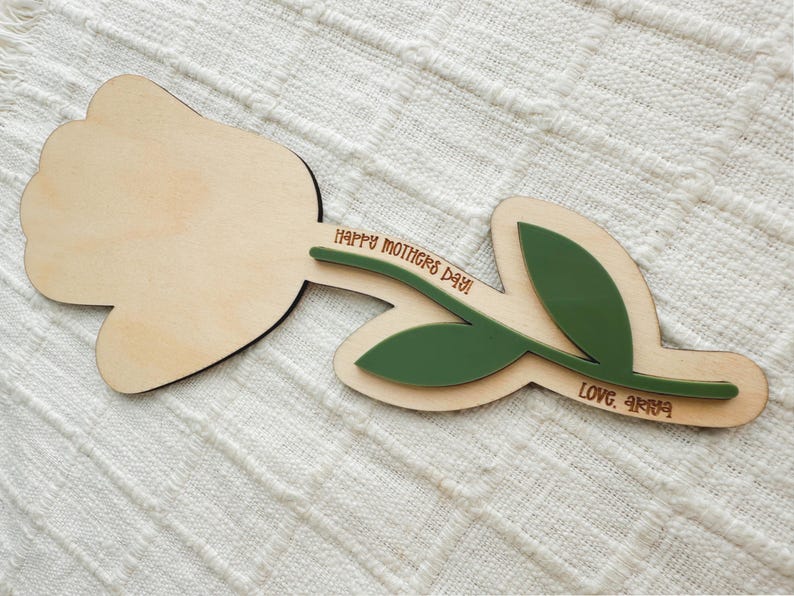 K&ouml;nnte beinhalten: Holzdekoration in Tulpenform mit hellem Holzfinish. Der Stiel und die Bl&auml;tter sind gr&uuml;n. Der Text "HAPPY MOTHERS DAY!" und "LOVE, ARYA" sind auf dem Stiel eingraviert. Die Dekoration liegt auf einer wei&szlig;en, strukturierten Oberfl&auml;che.