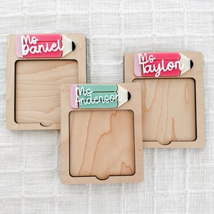 May include: Three wooden frames with a pencil design and a name on each. The names are Ms. Daniel, Ms. Taylor, and Ms. Anderson. The frames are light brown and have a white background.