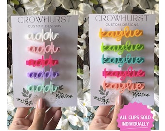 May include: Two sets of colorful hair clips with names spelled out. The first set spells out "addi" in pink, red, purple, and green. The second set spells out "kaylie" in yellow, green, blue, and pink. The text "ALL CLIPS SOLD INDIVIDUALLY" is on a pink starburst.