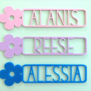 May include: Three personalised name plaques with a flower design. The top plaque is pink with the name "ALANIS", the middle plaque is light purple with the name "REESE", and the bottom plaque is blue with the name "ALESSIA".
