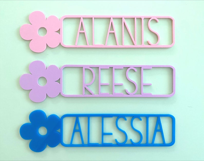May include: Three personalised name plaques with a flower design. The top plaque is pink with the name "ALANIS", the middle plaque is light purple with the name "REESE", and the bottom plaque is blue with the name "ALESSIA".