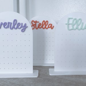 May include: Three white earring display stands with the names "Everley", "Stella", and "Ellie" written in different colours on top. Each stand has a grid of holes for holding earrings.