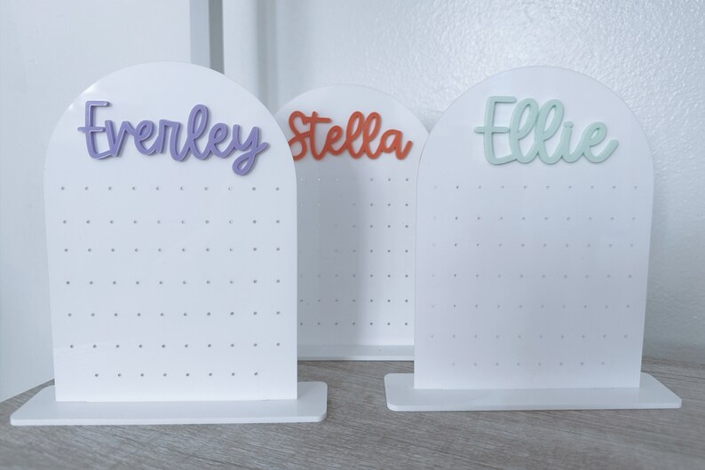 May include: Three white earring display stands with the names "Everley", "Stella", and "Ellie" written in different colours on top. Each stand has a grid of holes for holding earrings.