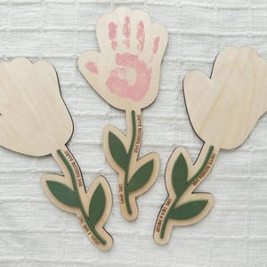 K&ouml;nnte beinhalten: Holzblumen-Ausschnitte mit gr&uuml;nen Stielen und Bl&auml;ttern. Eine Blume hat einen rosa Handabdruck. Die Stiele tragen die Aufschrift "Happy Mother's Day" und "Love you forever".