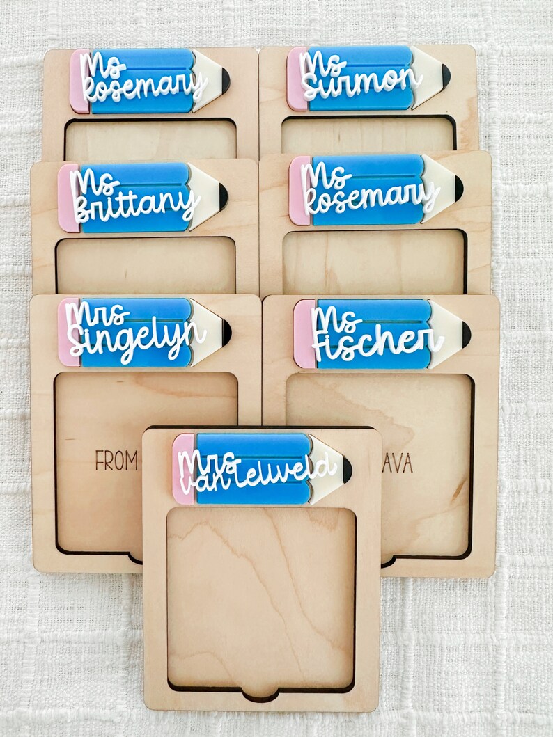 May include: Set of six wooden frames with a pencil-shaped cutout. Each frame has a different teacher's name written on it in white lettering. The frames are arranged in two rows of three. The top row reads "Ms. Rosemary", "Ms. Surmon", and "Ms. Brittany". The bottom row reads "Mrs. Singelyn", "Ms. Fischer", and "Mrs. VanTelweld".