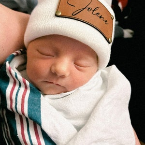 May include: A white knitted baby hat with a brown leather patch that says "Jolene" in cursive.