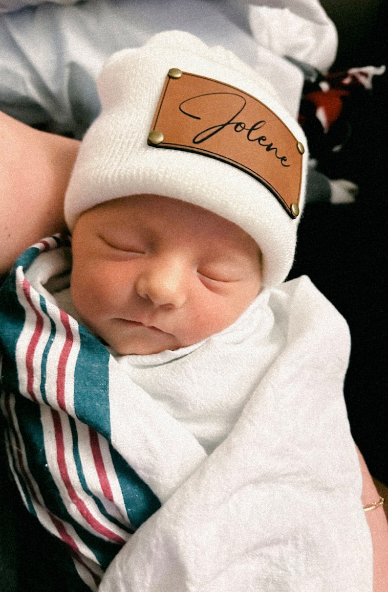 May include: A white knitted baby hat with a brown leather patch that says "Jolene" in cursive.