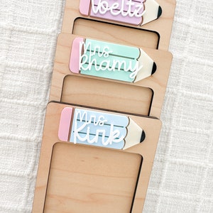 May include: Three wooden frames with pencil-shaped cutouts. Each frame has a different colored pencil with a pink eraser and white text that reads "Mrs. Voelz", "Mrs. Khamv", and "Mrs. Kirk".