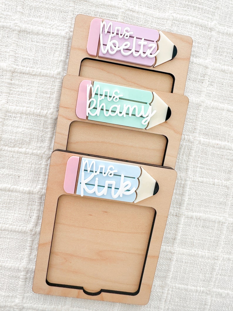 May include: Three wooden frames with pencil-shaped cutouts. Each frame has a different colored pencil with a pink eraser and white text that reads "Mrs. Voelz", "Mrs. Khamv", and "Mrs. Kirk".