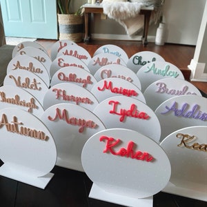 May include: A set of 16 white circular earring holders with different names written in various colours. Each holder has a small stand.