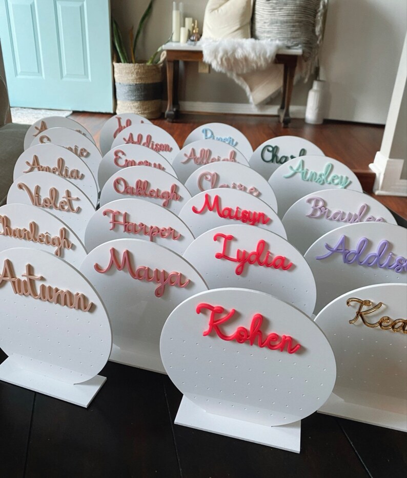 May include: A set of 16 white circular earring holders with different names written in various colours. Each holder has a small stand.