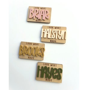 May include: Four wooden name plaques with the text "Look What Made" and a name in a contrasting color. The names are Briar, Halstyn, Brooks, and Hayes. The background is a light wood color.