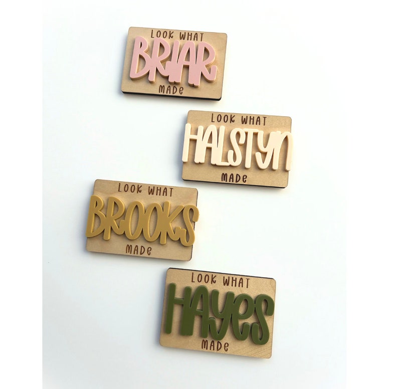 May include: Four wooden name plaques with the text "Look What Made" and a name in a contrasting color. The names are Briar, Halstyn, Brooks, and Hayes. The background is a light wood color.