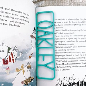 May include: A turquoise blue bookmark with the word "OAKLEY" printed in white letters. The bookmark is placed on a page of a book with illustrations of cartoon characters.