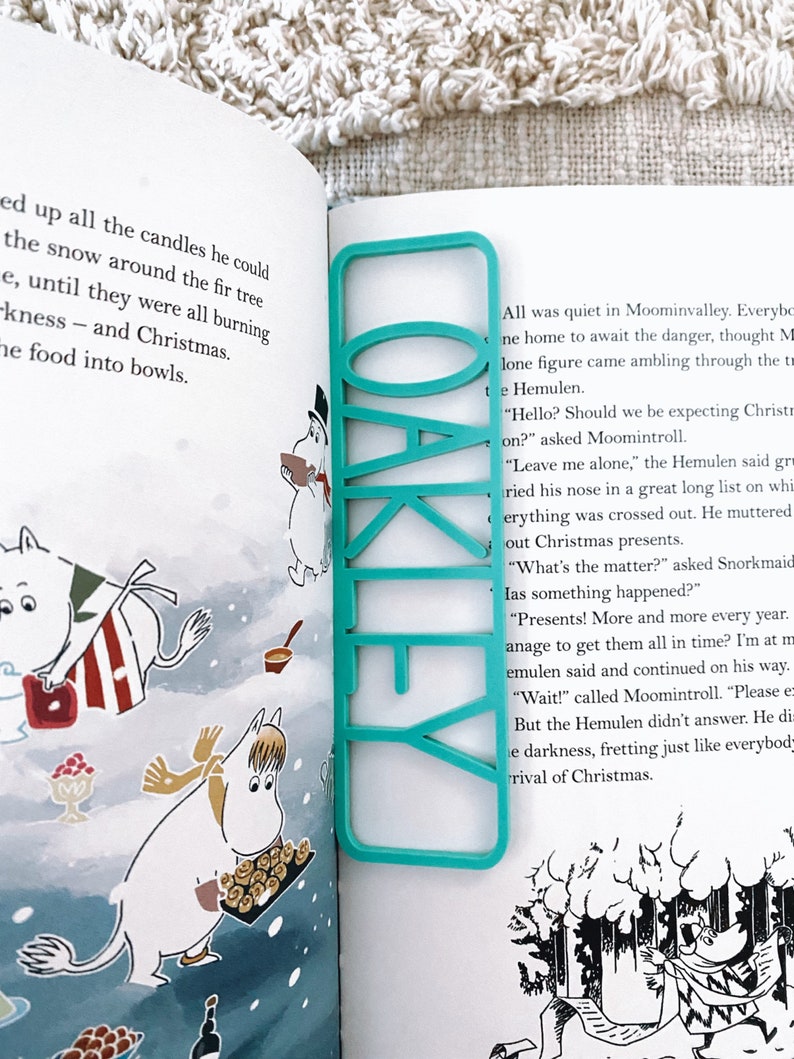 May include: A turquoise blue bookmark with the word "OAKLEY" printed in white letters. The bookmark is placed on a page of a book with illustrations of cartoon characters.
