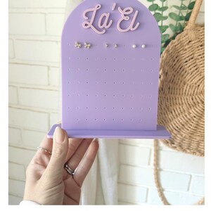 May include: A lilac earring display stand with the text "La' El" in a cursive font. The stand has multiple holes for earrings and is holding several pairs of earrings. The text "Ube on Lavender" is also visible.