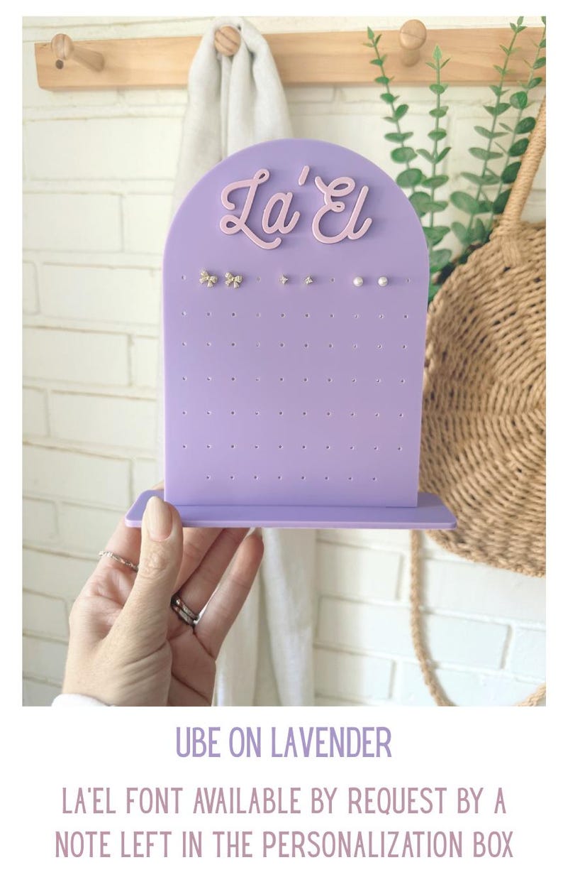 May include: A lilac earring display stand with the text "La' El" in a cursive font. The stand has multiple holes for earrings and is holding several pairs of earrings. The text "Ube on Lavender" is also visible.